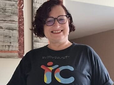 Mulher de pele clara, cabelos curtos e óculos de grau, com blusa preta com o logo da Tudo Sobre Inclusão e calça verde.